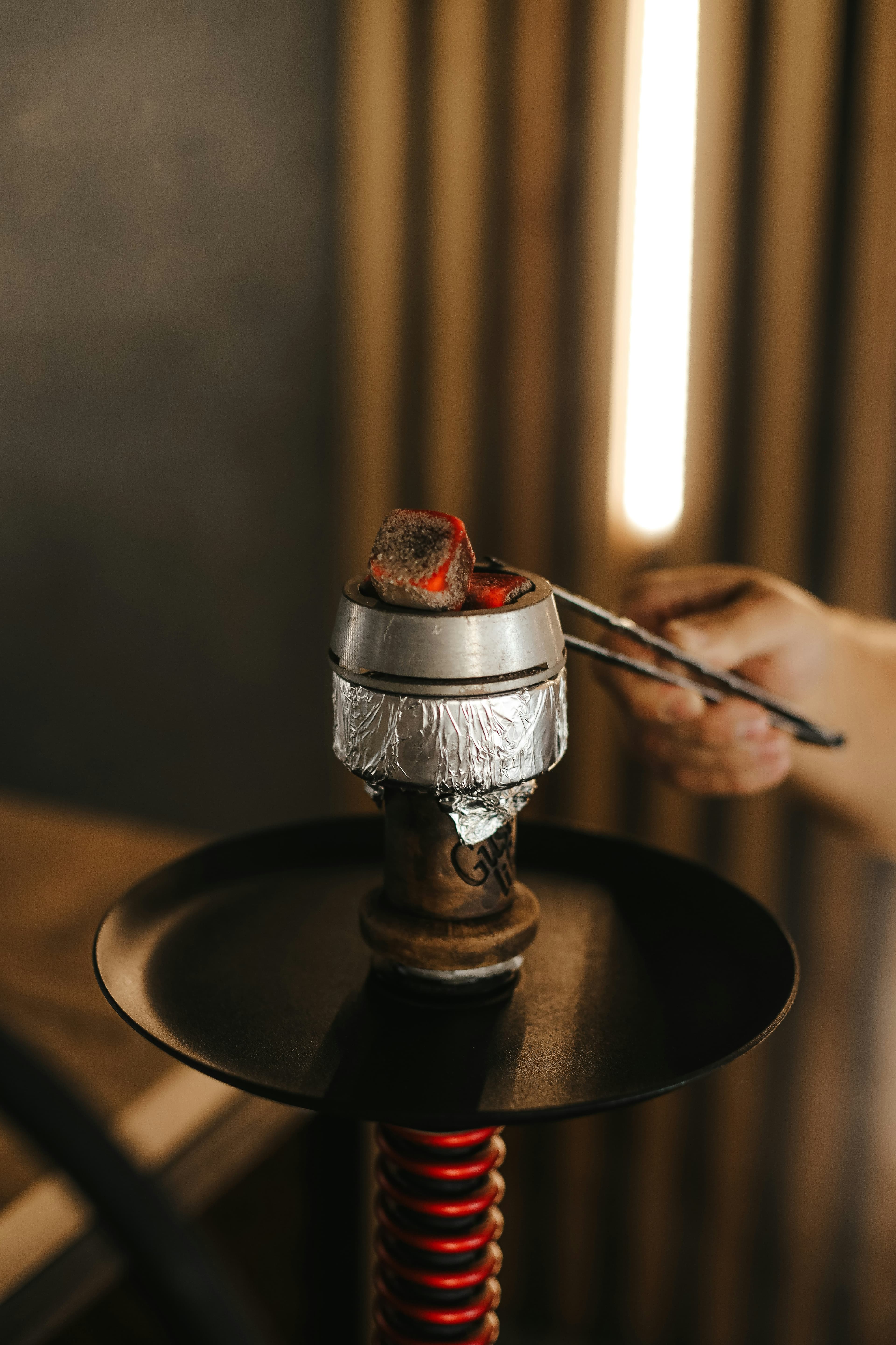 Hands placing a glowing coal with tongs during shisha preparation